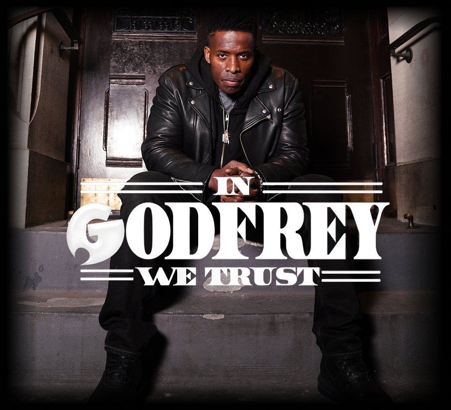 Godfrey sits on steps, leaning forward with hands clasped. He wears a black leather jacket and dark clothing, conveying a serious, contemplative mood.